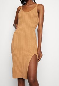 Kvinna som bär en ärmlös, ribbad, kamelfärgad stickad klänning med en hög lågspricka på ena sidan mot en vit bakgrund.