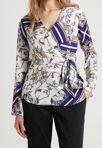 Top avvolgente in tessuto leggero con motivi floreali e geometrici in viola e bianco, con dettaglio a cravatta in vita e maniche lunghe.