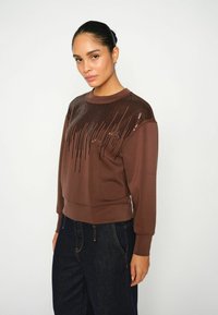 Hnedá mikina s textúrovaným dizajnom zdobeným flitrami na ramenách, so žebrovanými manžetami a lemom, skombinovaná s tmavými džínsami.