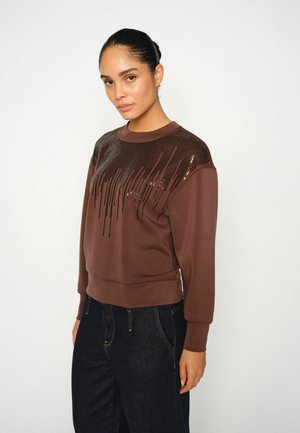 Brun sweatshirt med et struktureret, perlebesat design på skuldrene, ribstrikkede manchetter og kant, parret med mørke denimjeans.