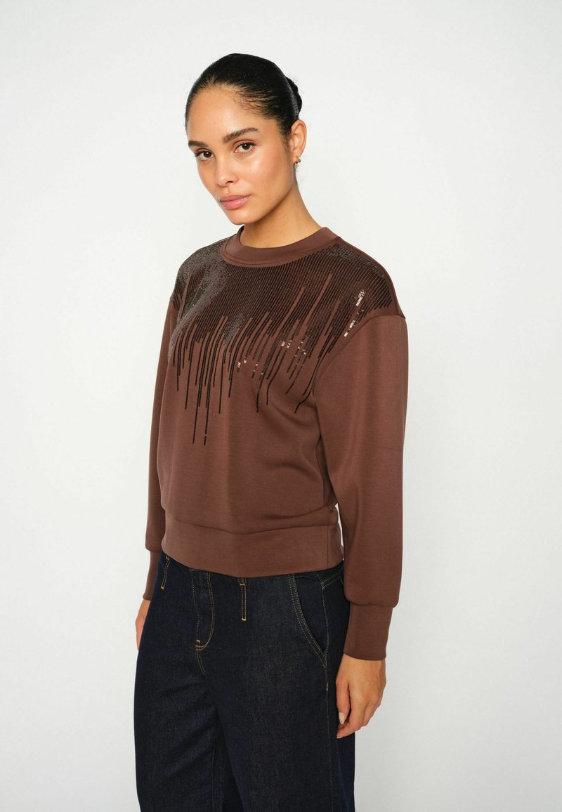 Barna pulóver, textúrált, flitterekkel díszített designnal a vállakon, bordás mandzsettákkal és aljjal, sötét farmernadrággal párosítva.