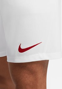 Shorts de sport blancs en tissu lisse, présentant un logo Nike rouge sur la jambe gauche. Le design est simple avec des ourlets cousus.