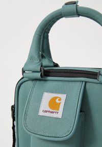 Morska platnena torba sa strukturnim dizajnom, gornjom ručkom, crnim patentnim zatvaračem i istaknutim Carhartt logotipom na bijeloj oznaci.