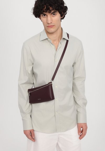 Geantă crossbody din piele burgundă, cu un fermoar argintiu și curea detașabilă, asortată cu o cămașă butonată de culoare gri deschis. Design simplu, rectangular.