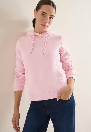 Rosa hoodie i mjukt tyg, med en framficka, justerbar huva med snören och ribbade fållar och ärmslut. Kombinerad med mörka jeans.