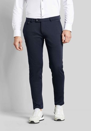 Mann trägt eine eng anliegende marineblaue Hose, weißes Hemd mit Knöpfen und weiße Turnschuhe und steht vor einem schlichten grauen Hintergrund.