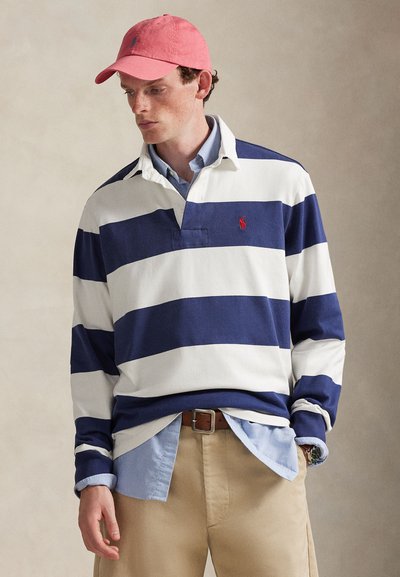 Chemise de rugby à manches longues rayée navy et blanc avec un logo rouge, superposée à une chemise à col bleu clair. Pantalon beige et casquette rose portés.
