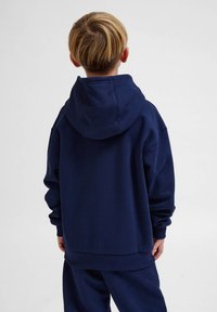 Mörkblå luvtröja i mjukt tyg, med en känguruficka och ribbade manschetter. Bärs av ett barn sedd bakifrån.