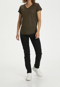 Olivově zelené tričko s krátkým rukávem a výstřihem do V v kombinaci s černými slim-fit kalhotami a bílými teniskami, na čistém bílém pozadí.