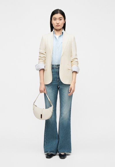 Γυναίκα που φοράει κρεμ σακάκι, γαλάζιο πουκάμισο, καμπανοειδές μπλε τζιν, μαύρα παπούτσια, κρατώντας λευκή τσάντα με χρυσό κούμπωμα μπροστά σε μονόχρωμο φόντο.