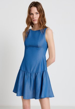 Giovane donna con capelli mossi che indossa un vestito di jeans blu senza maniche fino al ginocchio con gonna svasata, in piedi davanti a uno sfondo chiaro e semplice.