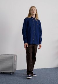 Camisa de mezclilla de botones en azul oscuro, pantalones beiges y zapatillas deportivas azules. El fondo incluye un contenedor de almacenamiento gris sobre una alfombra.
