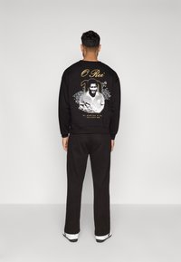 Čierna mikina s grafickou potlačou, ktorá na chrbte zobrazuje bielu a zlatú ilustráciu, a zodpovedajúce čierne tepláky.