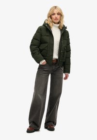 Chaqueta acolchada verde oscura con capucha, combinada con un suéter blanco de punto trenzado y jeans de pierna ancha grises. Incluye zapatos marrones y un cinturón trenzado.
