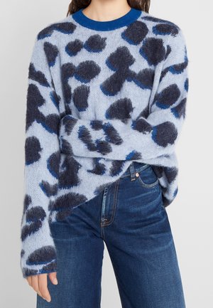 Kvinde iført en blød blå sweater med mørkeblå og sorte abstrakte pletter sammen med mørkeblå jeans.
