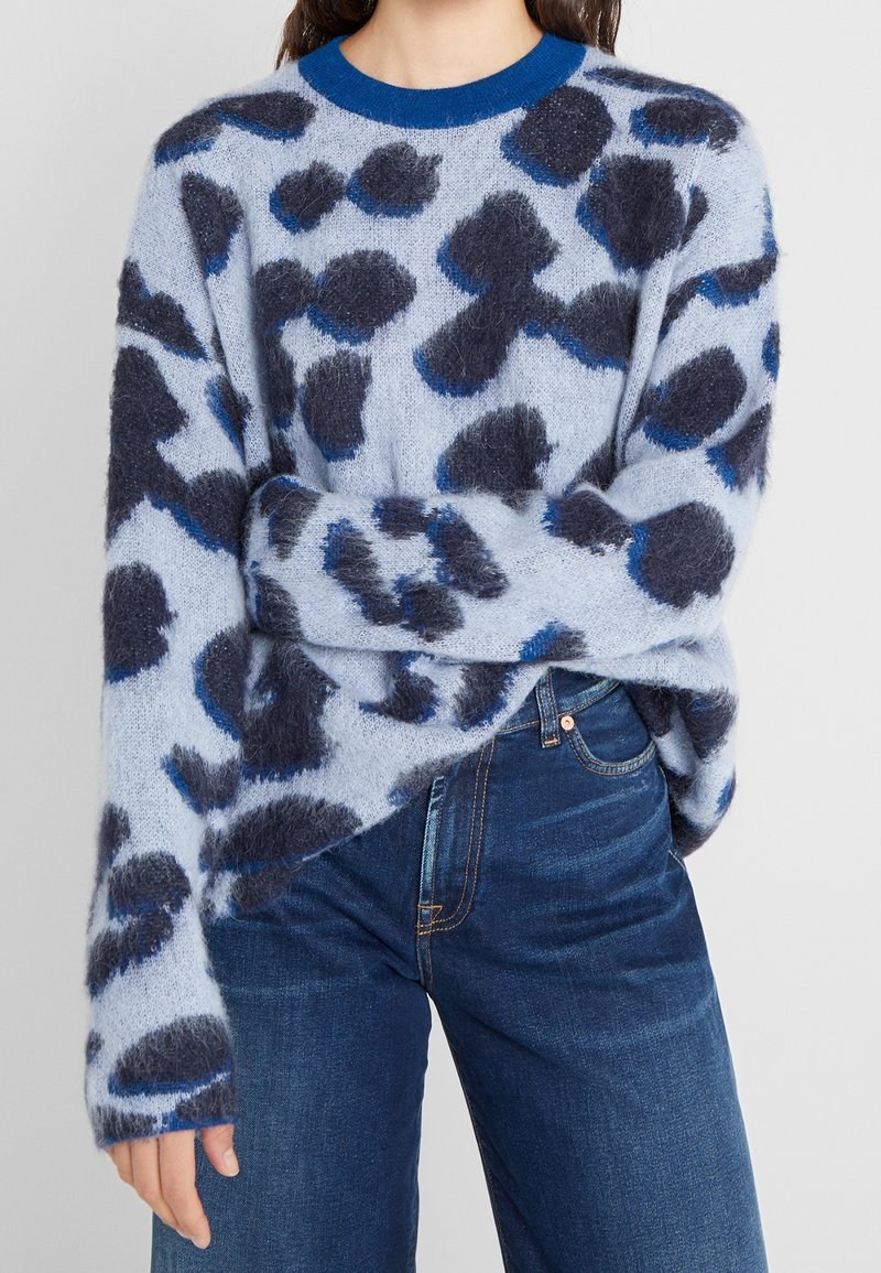 Femme portant un pull bleu duveteux avec des taches abstraites bleu foncé et noires, associé à un jean bleu foncé.