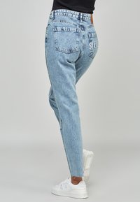 Světle modré vysoké mom džíny vyrobené z denimu, s volným střihem, pěti kapsami a potrhaným lemem. Bílé logo na zadní kapse.