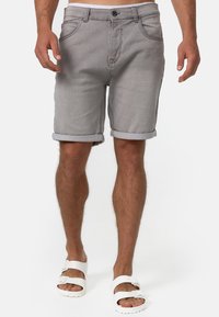 Shorts en denim gris avec des revers, comportant des poches avant et un bouton de fermeture, associés à des sandales blanches sur pieds nus.