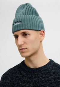 Strikket beanie i teal med lodret ribbede strukturer og broderet logo. Båret af en person sammen med en mørk, tekstureret sweater.