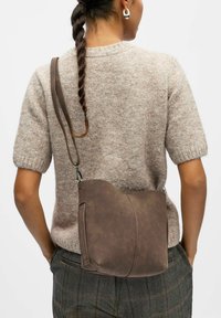 Femme avec des cheveux tressés portant un pull beige à manches courtes et un pantalon à carreaux, portant un sac en cuir marron en bandoulière sur le dos.