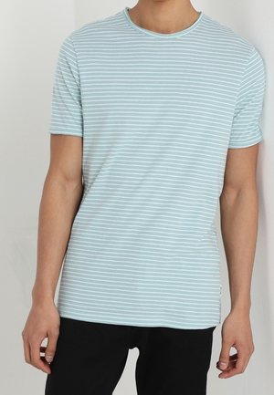 Man wearing light blue and white thin horizontally striped short-sleeve T-shirt and black pants, standing against a white wall.