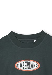 Tamnozelena pamučna majica s okruglim izrezom i grafičkim logotipom koji prikazuje "TIMBERLAND" u narančastoj i crnoj boji unutar ovala.