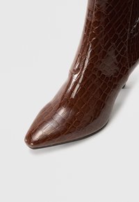 Botín de tobillo marrón con estampado de cocodrilo y acabado brillante, punta afilada y tacón delgado. El material parece ser sintético o imitación de cuero.