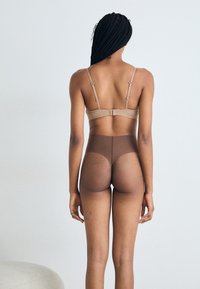 Beige bh med tynne stropper og høy midje gjennomsiktige brune strømpebukser, sett fra baksiden. Jevn tekstur med subtil fargevariasjon.