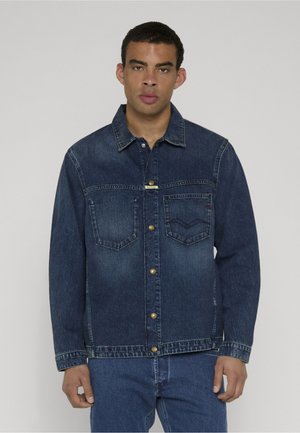 Young man wearing a dark blue denim jacket with metal buttons and front pockets, paired with blue denim jeans, standing against a plain background.