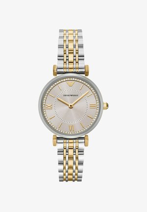 Dvoubarevné kovové hodinky se stříbrným a zlatým páskem. Kulatý stříbrný ciferník ozdobený krystaly. Římské číslice na 6 a 12 hodin, zlaté ručičky.