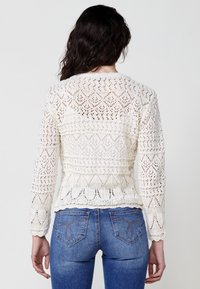 Bílý pletený svetr s texturovaným krajkovým vzorem, dlouhými rukávy a přiléhavým pasem. Spárován s modrými džínami z denimu s dekorativním prošitím.