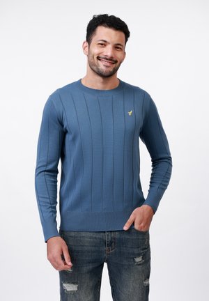 Suéter azul de canalé con mangas largas, cuello redondo y un acento de logo dorado en el pecho, combinado con jeans de mezclilla desgastados.