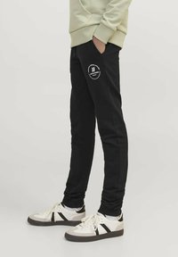 Pantalones de chándal negros de mezcla de algodón, con un diseño ajustado, bolsillos laterales y un logo impreso en el muslo izquierdo. Combinados con zapatillas blancas.