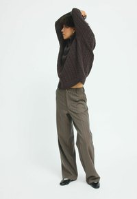 Pull en tricot torsadé marron au look surdimensionné avec col roulé, associé à un pantalon marron à rayures. Le mannequin se tient de profil dans un cadre minimaliste.