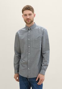 Sivi button-up košulja s oštrim ovratnikom, jednim džepom na prsima i dugim rukavima; izrađena od teksturirane tkanine. Nosena s plavim trapericama.