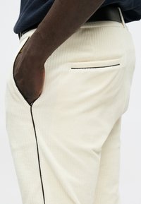 Kremowe spodnie o prążkowanej fakturze, z czarnym lamowaniem wzdłuż krawędzi kieszeni i luźnym krojem. Widoczna ręka w kieszeni.