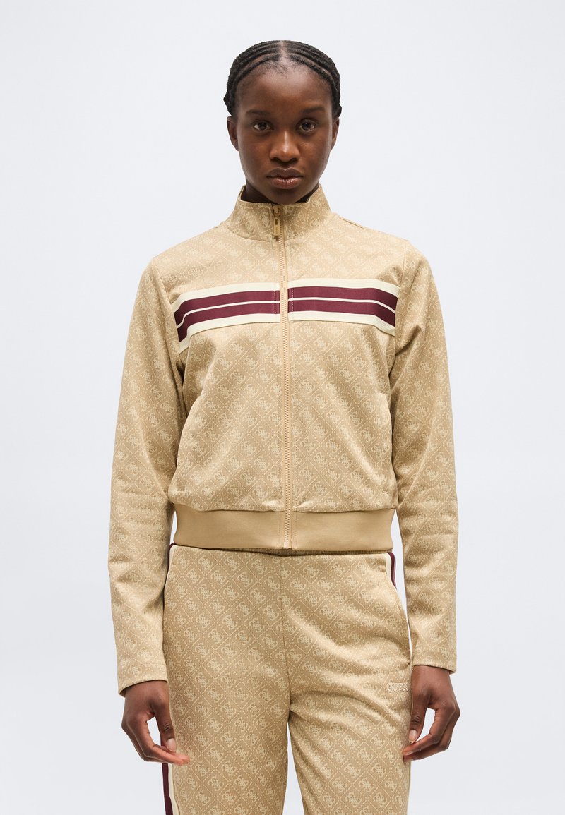 Persoon draagt een beige gestreepte jas met ritssluiting en bijpassende broek met bordeauxrode en witte strepen over de borst en langs de zijkanten.