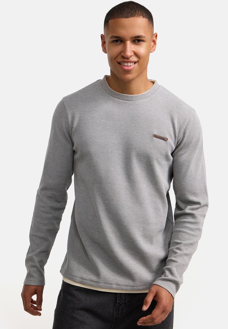Graues Langarmshirt mit einer strukturierten, gerippten Oberfläche. Besitzt einen runden Halsausschnitt und dezente Markenkennzeichnung auf der Brust. Wird mit dunklen Hosen kombiniert.
