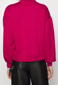 Fuksjowy sweatshirt z wysokim kołnierzem i bufiastymi rękawami, z prążkowanym dołem. W połączeniu z czarnymi spodniami cargo z ekoskóry.