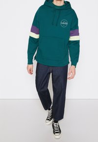 Teal hoodie med lila och ljusgula ränder, en frontficka och logotypdetalj. Kombinerad med mörka denimjeans och svarta sneakers.