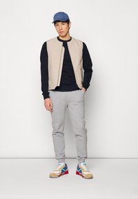 Beige mouwloos vest over een zwart langemouwen shirt, grijze joggingbroek en meerkleurige sneakers. Blauwe pet op het hoofd. Eenvoudige achtergrond.