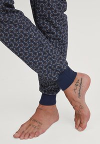 Marine joggers met een herhalend regenboogpatroon, elastische manchetten en een geribbelde tailleband. Blote voet zichtbaar met meerdere tatoeages.