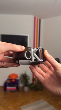 Cintura in pelle marrone con fibbia color argento con le lettere "CK". La cintura è tenuta tra due mani, mostrando la sua texture liscia.