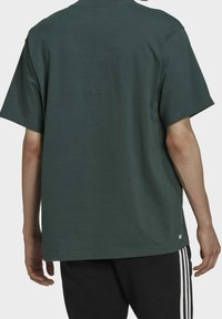 Eine Person trägt ein dunkelgrünes Kurzarm-T-Shirt und schwarze Hosen mit weißen seitlichen Streifen und steht mit dem Rücken zur Kamera.