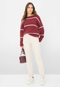Burgunderfarbener Strickpullover mit weißen Streifen, kombiniert mit beigen Hosen und weißen Turnschuhen. Das Model hält eine kleine burgunderfarbene Handtasche.