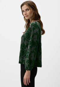 Blusa con motivo floreale verde a maniche lunghe, realizzata in un tessuto texturizzato. Presenta una vestibilità rilassata e polsini elastici, abbinata a pantaloni neri.