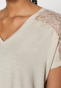 Jasnoszara bluzka z krótkim rękawem, z dekoltem w kształcie litery V i koronkowym wykończeniem na ramionach, wykonana z miękkiego materiału o lekko teksturowanym wykończeniu.