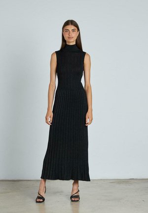 Schwarzes geripptes Maxikleid mit hohem Ausschnitt, tailliertem Oberteil und weit ausgestelltendem Saum. Getragen mit schwarzen Riemchensandalen. Glatte Textur.
