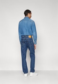 Camisa de mezclilla en azul claro, de mangas largas, con puños abotonados; combinada con jeans de mezclilla azul oscuro, con bolsillos traseros visibles, etiqueta de cuero en la cintura.