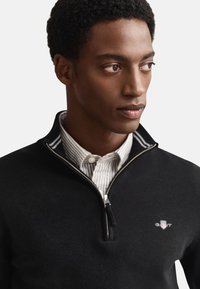 Crni džemper s patentnim zatvaračem i visokim ovratnikom, s malim logom na prsima i prugastim detaljima na ovratniku. Mekana tekstura.